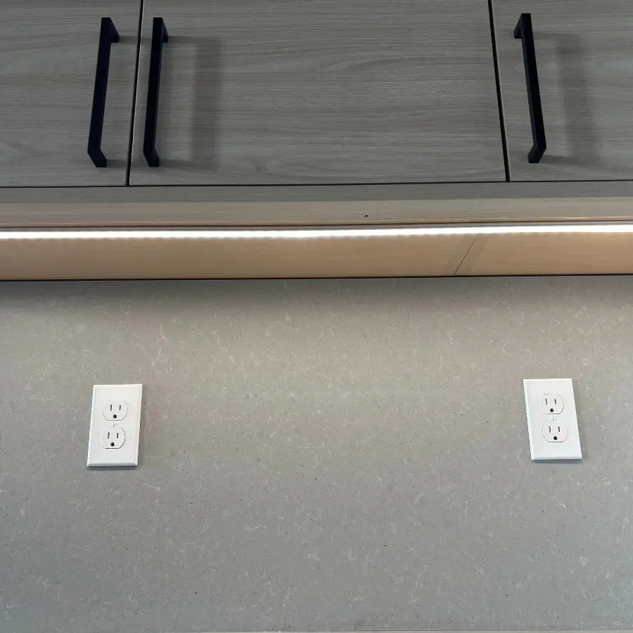A modern kitchen with light gray wooden cabinets and sleek black handles. Under the cabinets, a strip of LED lighting illuminates a light gray countertop.